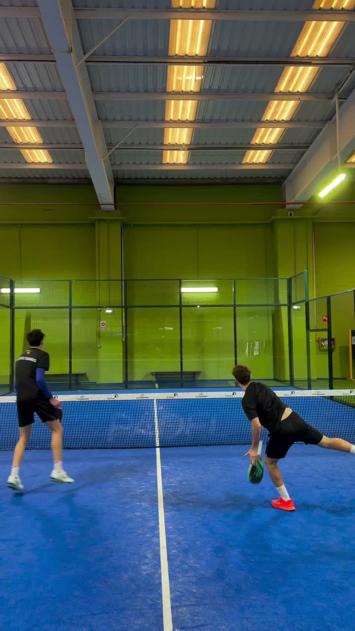 Two players on a blue indoor pickleball/tennis court near a net, one in black bending to hit a green paddle while the other watches.