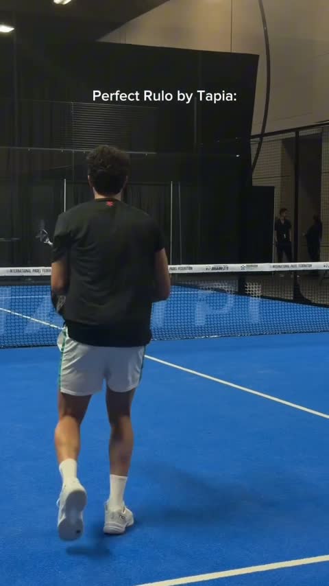 Back view of a man walking on a blue indoor tennis court toward the net, wearing a black shirt and white shorts.
